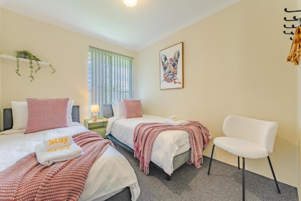 Habitación de hotel con dos camas, una silla blanca, una ventana con persianas, un cuadro en la pared y una toalla con la palabra 'Stay'.