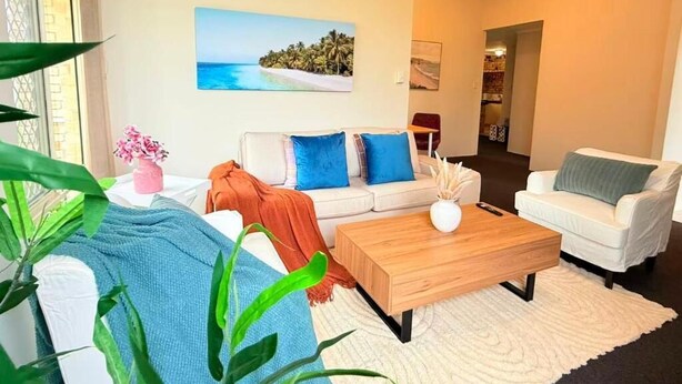 A living room with a white sofa, blue cushions, a wooden coffee table, and a painting of a beach scene. - StayAU 4BR Family Retreat with Garden (Seville Grove)
