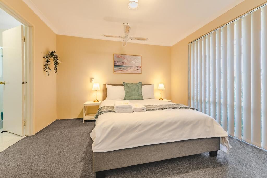 A bedroom with a bed, two bedside lamps, a ceiling fan, a painting on the wall, and a window with blinds.