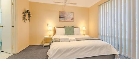 A bedroom with a bed, two bedside lamps, a ceiling fan, a painting on the wall, and a window with blinds.