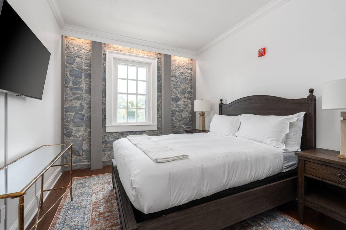 Une chambre dotée d’un grand lit, d’un téléviseur à écran plat fixé au mur, d’un mur décoratif en pierre et d’une fenêtre donnant sur un paysage verdoyant.