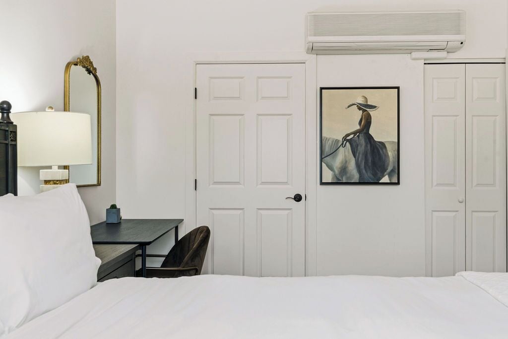 A hotel room with a bed, a desk with a lamp, a chair, a large mirror, and a door with a painting.