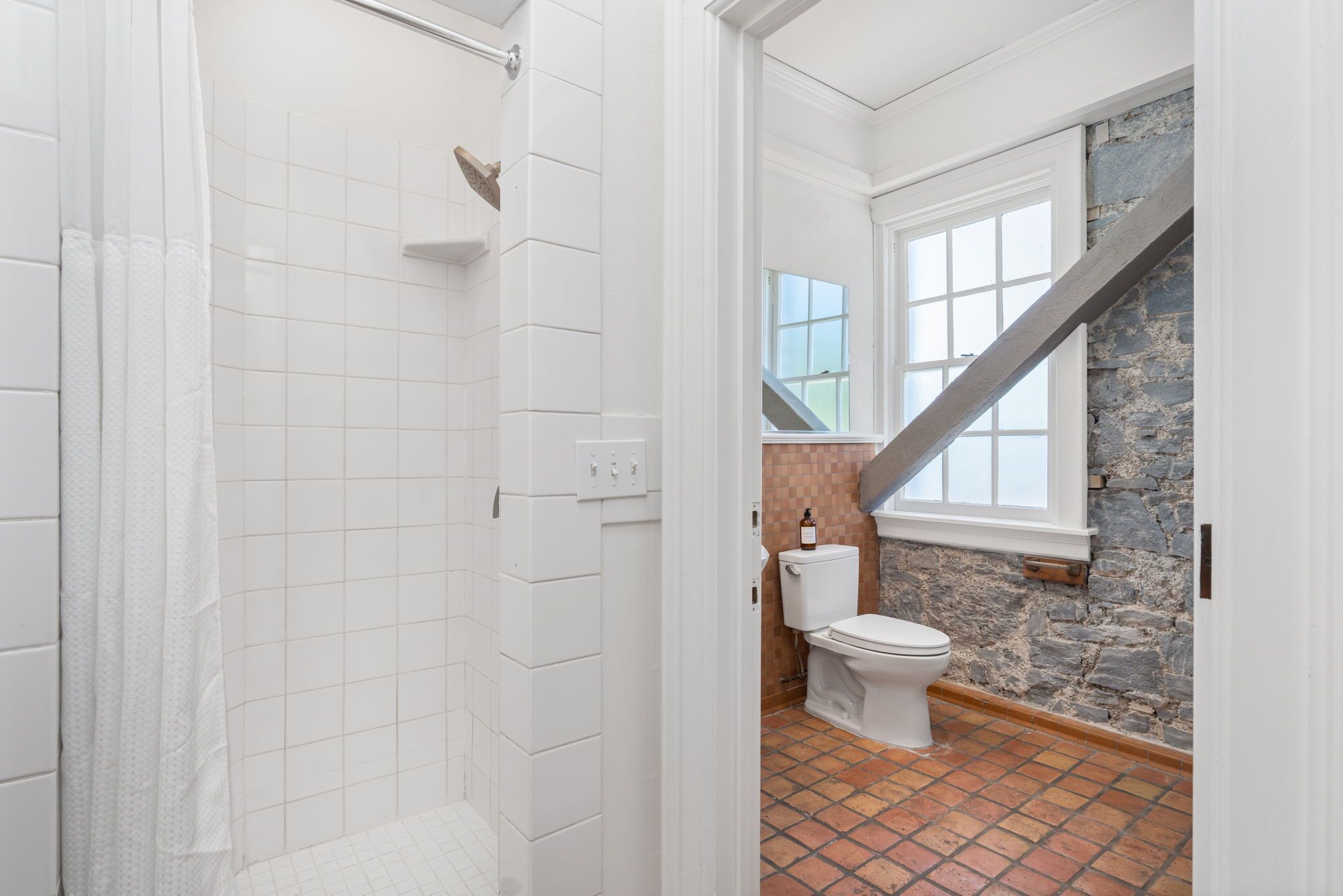 Une salle de bain avec des murs recouverts de carreaux blancs, une douche, des toilettes et un mur d’accent en pierre.