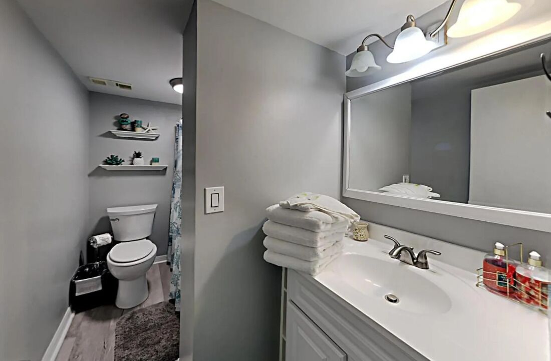 Un baño moderno con inodoro, lavamanos y espejo. Hay repisas con objetos decorativos sobre el inodoro y una pila de toallas al lado del lavamanos.