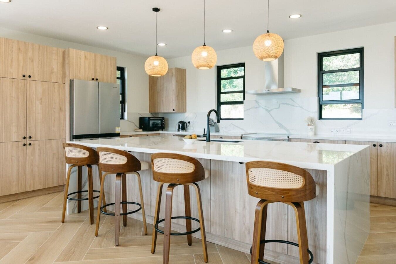 Una cocina moderna con una isla central, taburetes de madera, iluminación colgante y un ventanal amplio.