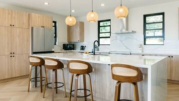 Una cocina moderna con una isla central, taburetes de madera, iluminación colgante y un ventanal amplio.