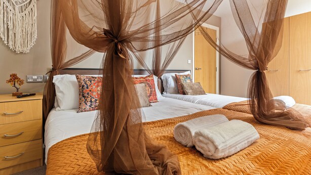 Uma cama de casal com dossel marrom, um criado-mudo de madeira e um painel de parede em macramé, além de um guarda-roupa de madeira.
