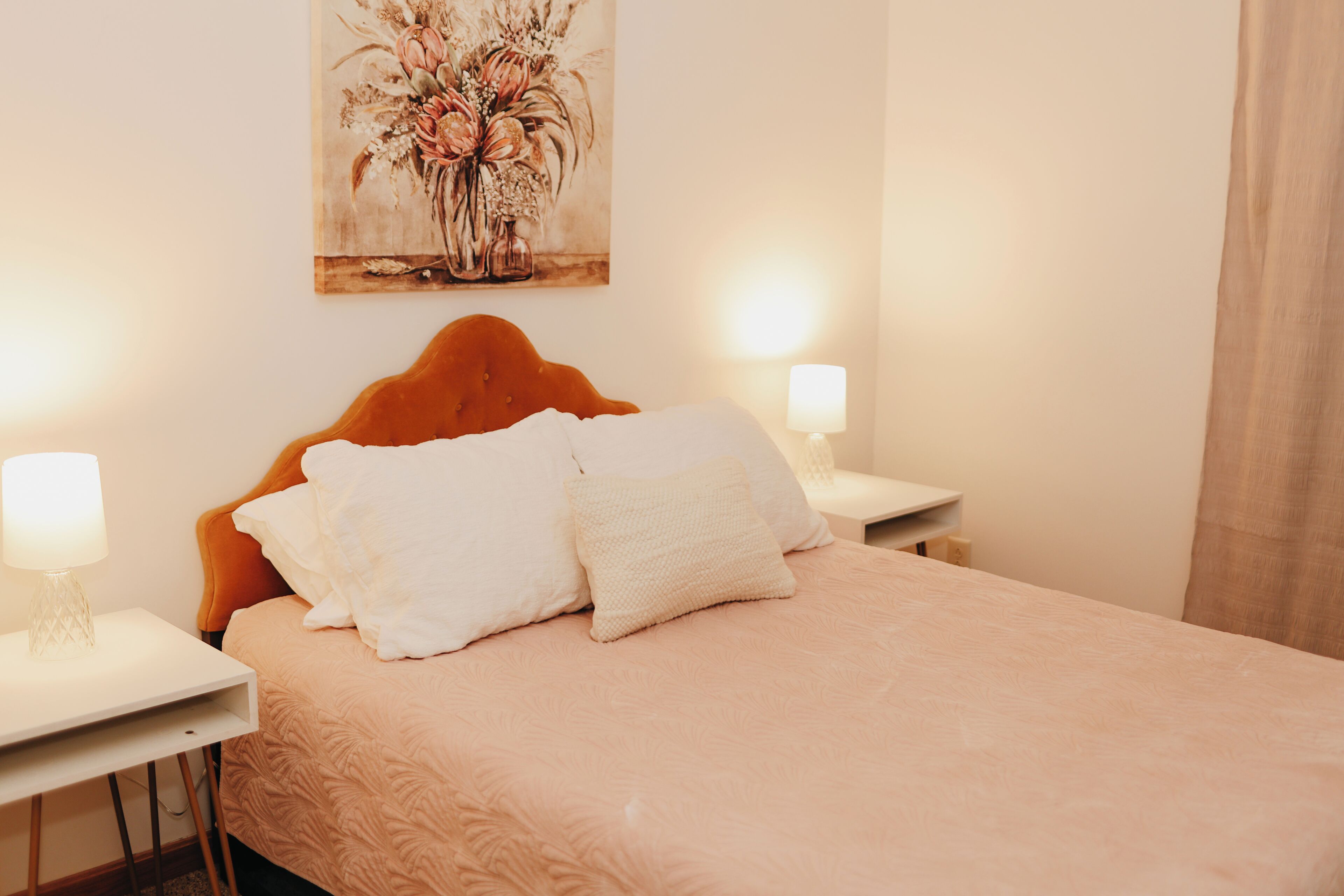 A neatly made bed with a headboard, two bedside tables with lamps, and a floral painting on the wall.