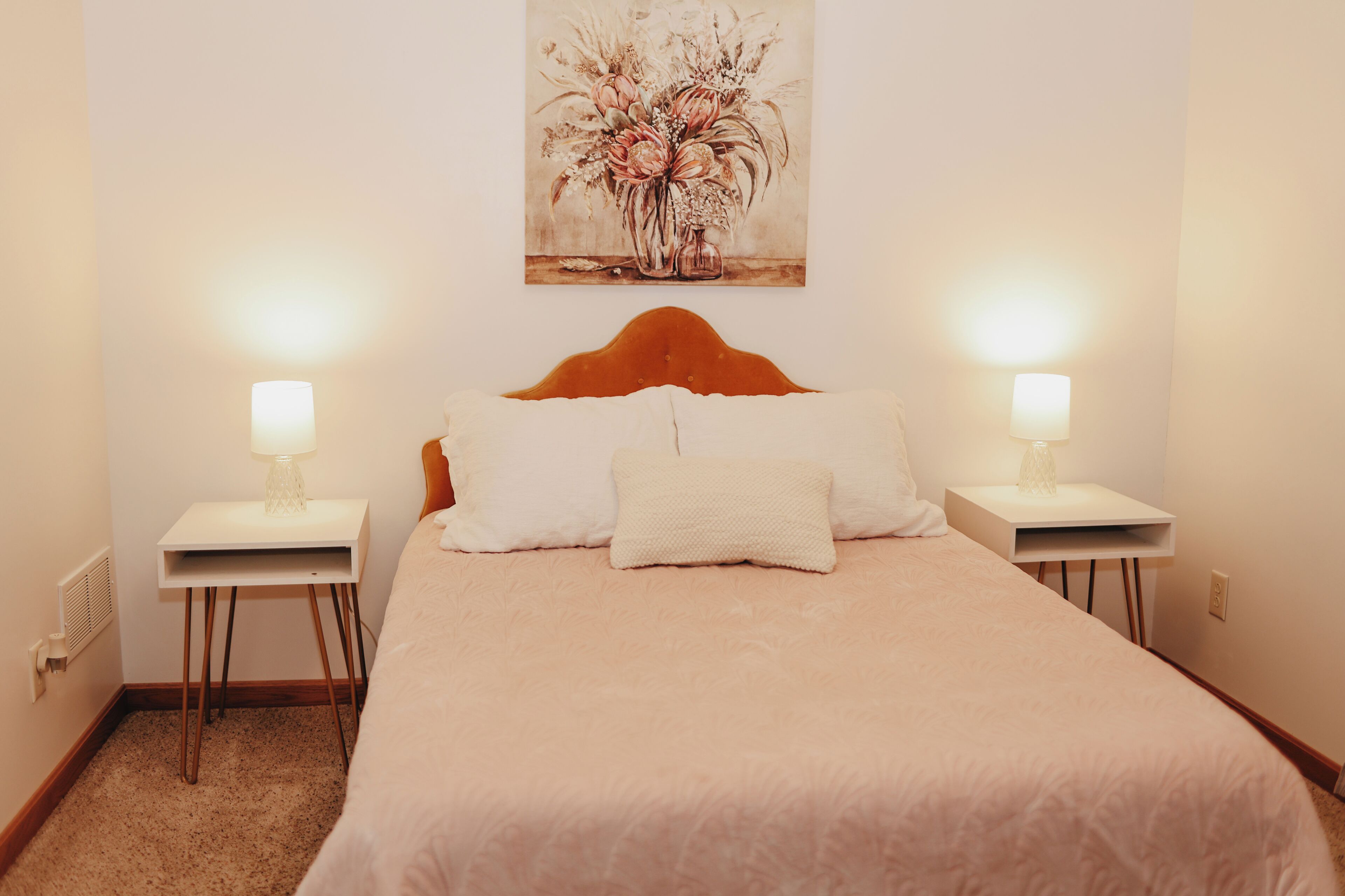 Un dormitorio con una cama grande, dos mesitas de noche con lámparas, un cabecero y un cuadro con motivos florales en la pared.