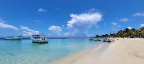Uma praia com águas turquesa cristalinas, vários barcos e uma faixa de areia bordeada por palmeiras.