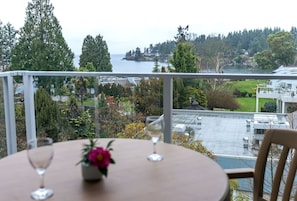 A round table with two wine glasses and a flower vase, overlooking a view of a body of water, trees, and buildings.