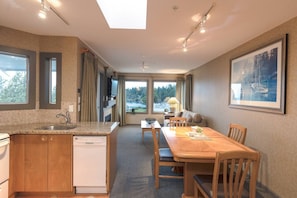 A hotel room with a kitchen area, a dining table with chairs, a sofa, and a large window with a view.