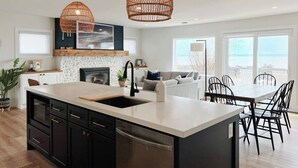 A modern kitchen with a large island, black cabinets, and a built-in dishwasher. There is a dining area with a table and chairs, and a sofa. The kitchen has a white backsplash and a large window overlooking the ocean. - Lakefront Cottage + Suite  | 4 Bedrooms | 2 Connected Cottages Family Retrea (Vittoria)