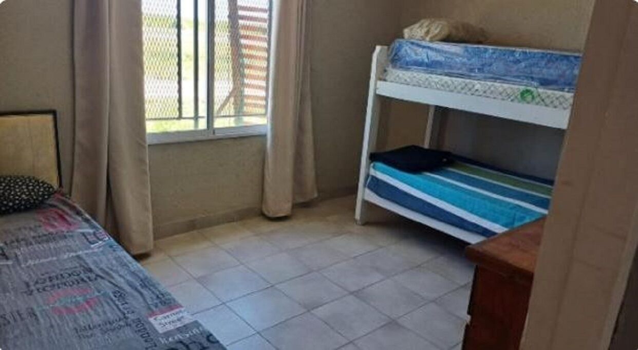 A small room with a bunk bed, a single bed, a window with curtains, and a tiled floor.