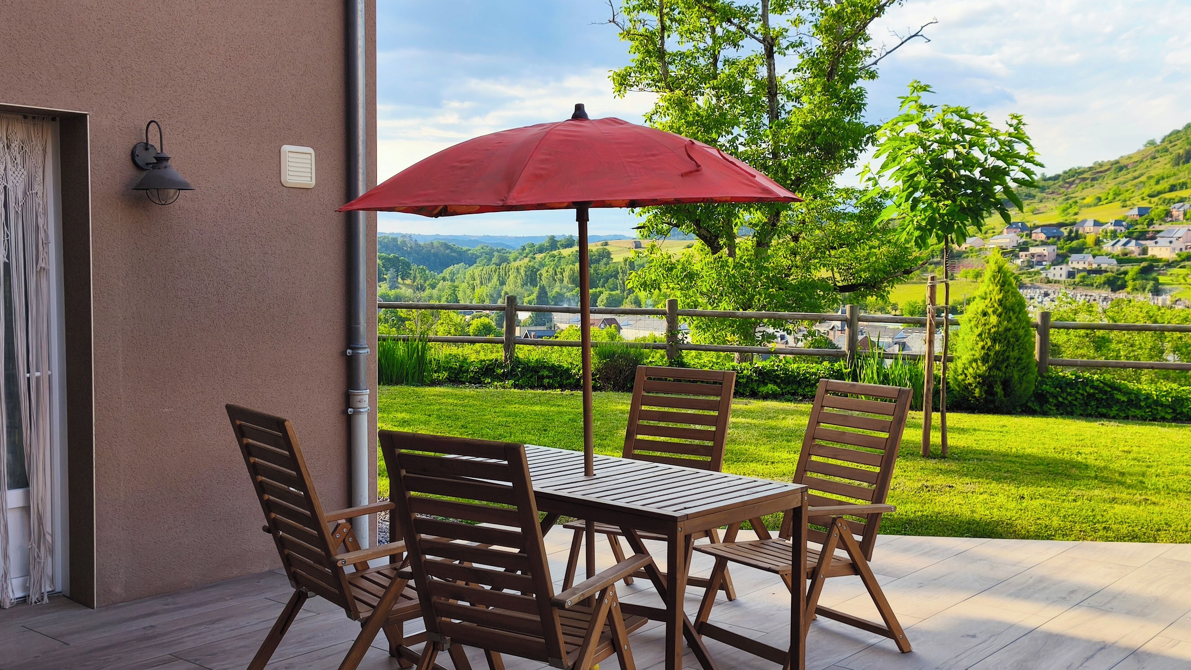 Eine Terrasse mit Tisch und Stühlen, einem roten Sonnenschirm und Blick auf eine grüne Landschaft mit Häusern in der Ferne.