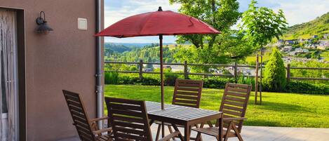 Eine Terrasse mit Tisch und Stühlen, einem roten Sonnenschirm und Blick auf eine grüne Landschaft mit Häusern in der Ferne.