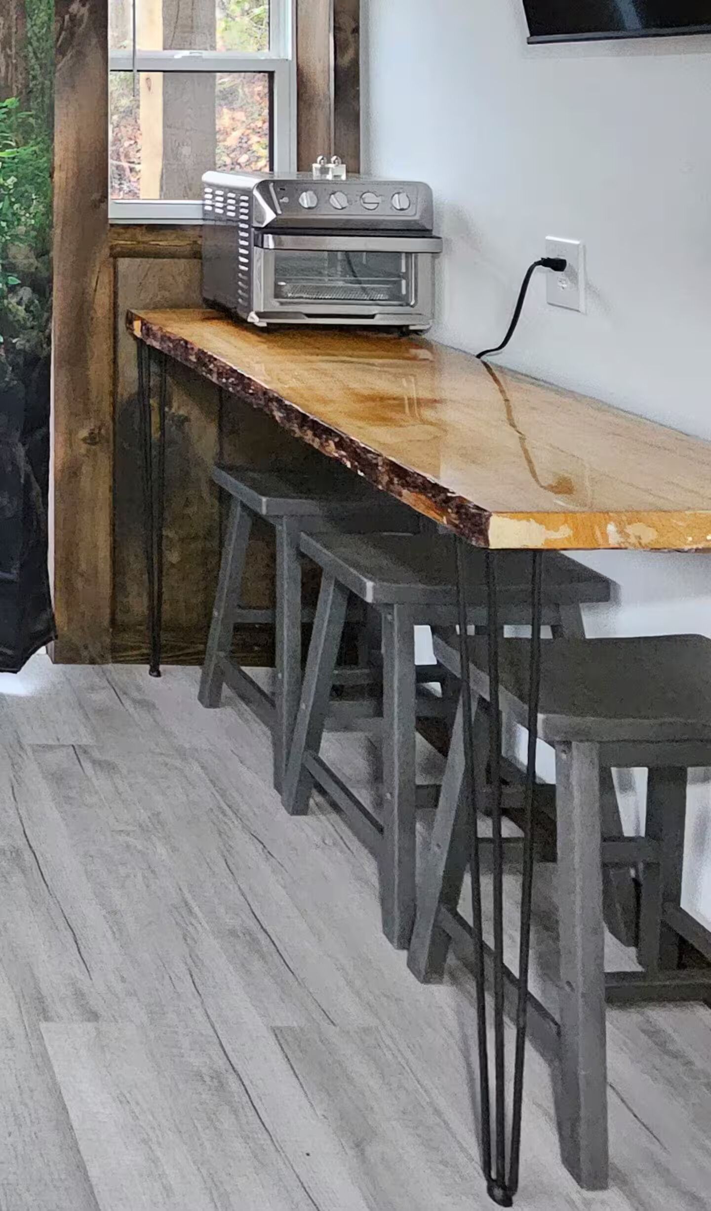 A rustic kitchen with a wooden bar counter and stools, a built-in oven, and a window with a view of trees.