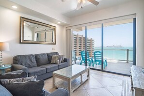 A modern living room with a grey sofa, a glass coffee table, and a view of the sea through large sliding doors.