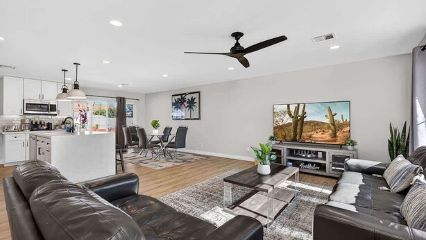 Una sala de estar moderna con un sofá, una mesa de centro y un televisor. Se observa una cocina al fondo con un comedor.