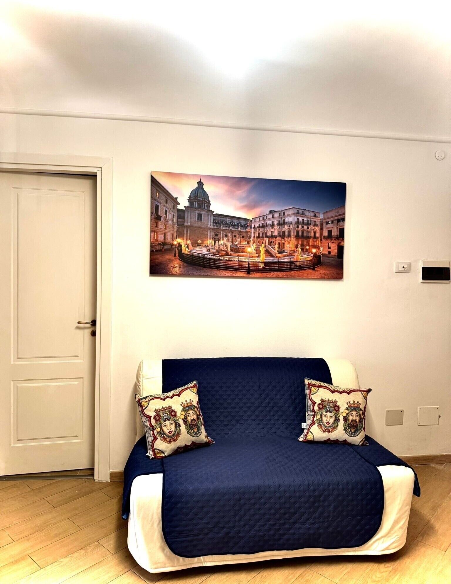 A bedroom with a bed, two pillows, a wall-mounted painting of a cityscape, and a door.