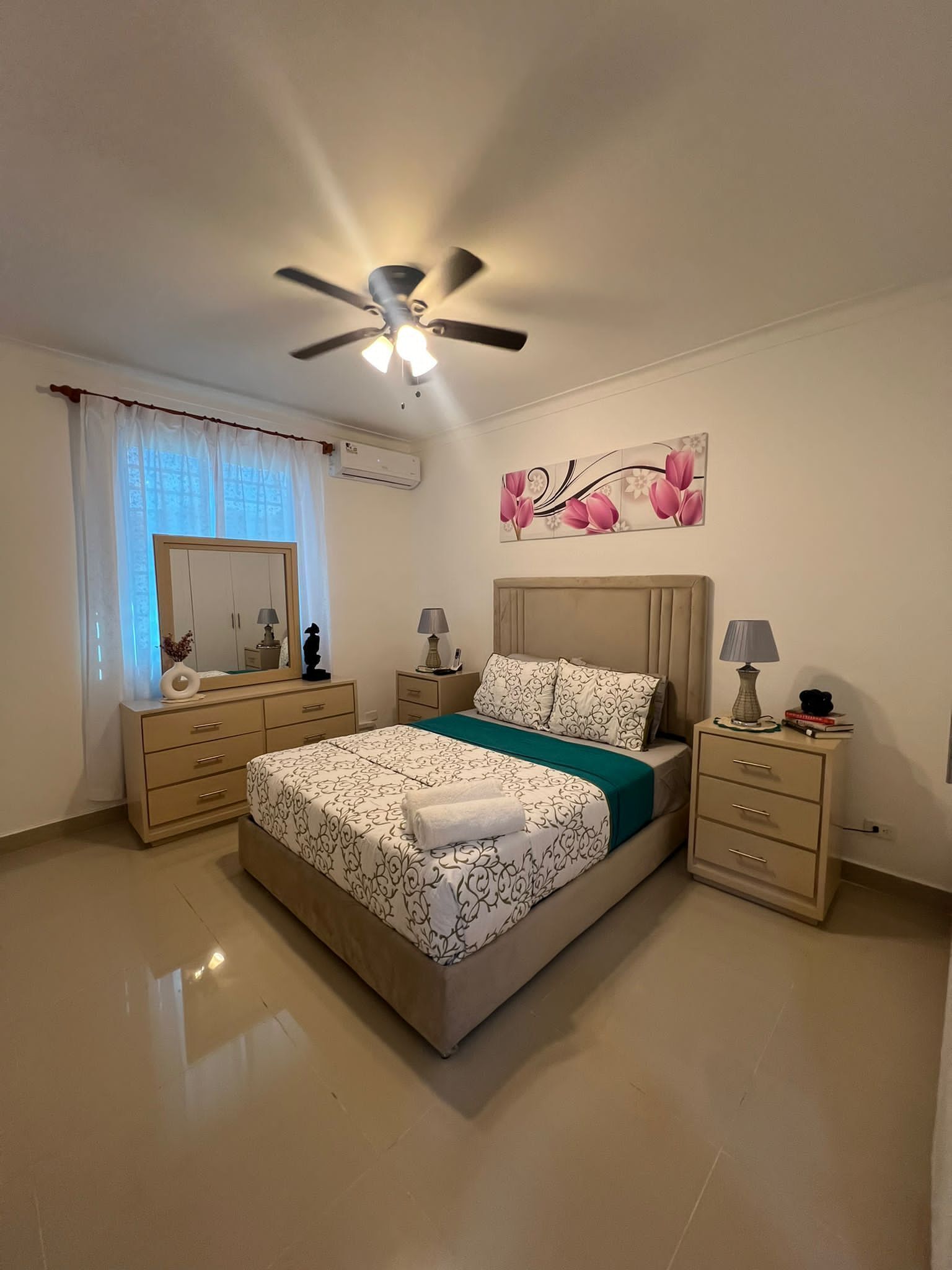 Ein Schlafzimmer mit einem Bett, Nachttischen, einem Deckenventilator, einem Fenster mit Vorhängen und einer Wand-Klimaanlage.