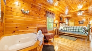 Ein Holzblockhaus-Interieur mit einer großen Badewanne, einem Bett mit Karo-Bettwäsche und einem kleinen Tisch mit einer Lampe.