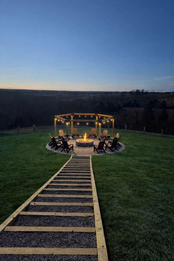 A fire pit area with seating and a wooden deck leading to it, surrounded by a grassy field and a clear sky.