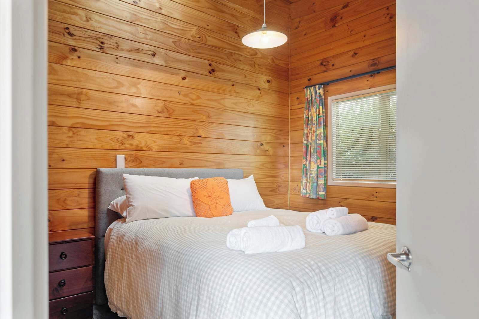 A wooden-paneled bedroom with a bed, pillows, a dresser, and a window with blinds.