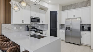 Una cocina moderna con armarios blancos, una isla central con pileta, electrodomésticos de acero inoxidable e iluminación colgante.