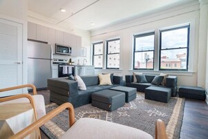 A modern living room with a grey sectional sofa, a small kitchen area, and large windows offering city views.