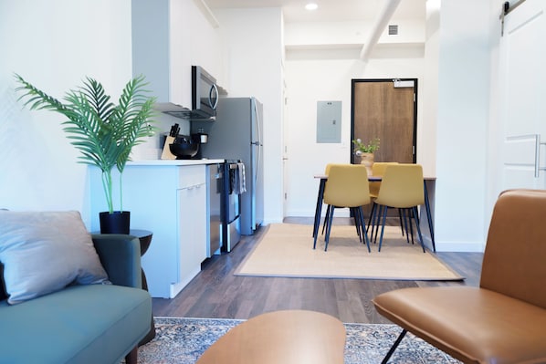 A modern kitchen with a refrigerator, microwave, and stove. There is a dining area with yellow chairs and a table. A sofa and a potted plant are also visible. - The Standard in Downtown Columbus (Columbus)