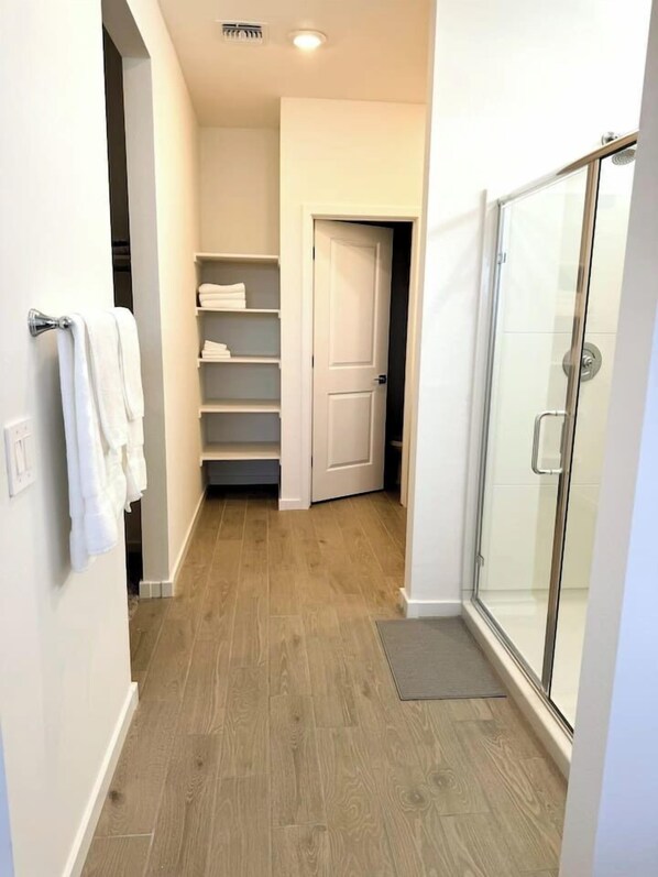 A narrow hallway with a wooden floor, a glass shower enclosure, and a white door leading to a closet with shelves. - New 4BR Casa Grande Home | Pool, Golf + Skydive AZ (Casa Grande)