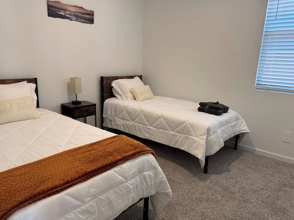 A bedroom with two beds, a nightstand, a lamp, and a window with blinds. - Modern 3BR Casa Grande Home | Pool Access + I-10 (Casa Grande)