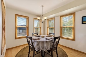 A dining room with a round table, four chairs, a chandelier, and large windows with blinds. - Ada O’Day Condominium #2 – Lake Superior Views in the Heart of Bayfield (Bayfield)