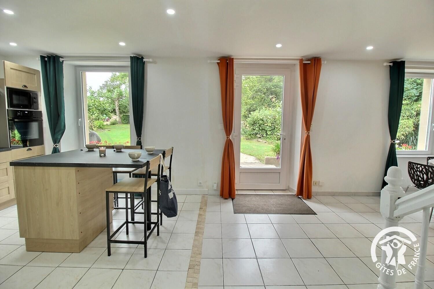 A modern kitchen with a central island, dining table, and chairs. There is a built-in oven and a view of the garden through the windows.