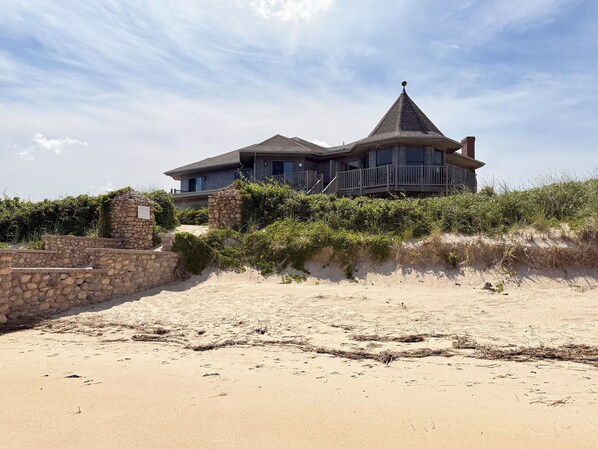 A beachfront house with a stone wall, a wooden deck, and a sandy beach in front. - Linger Longer by the Sea (Brewster)