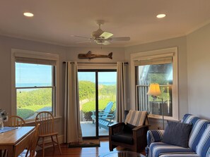 A room with a dining table, chairs, a sofa, a ceiling fan, and large windows with a view of greenery and the sea. - Linger Longer by the Sea (Brewster)