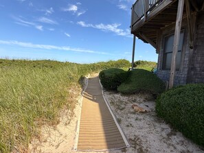 A wooden pathway leading to a building with a balcony, surrounded by sand and vegetation. - Linger Longer by the Sea (Brewster)