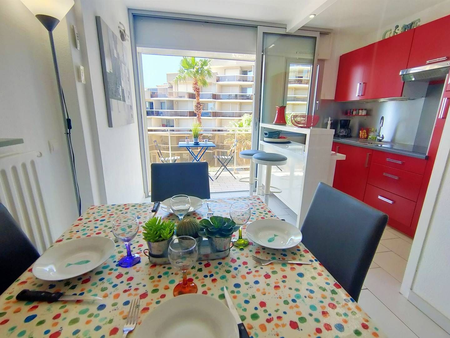 A dining area with a table set for two, a kitchen with red cabinets, and a view of a balcony with outdoor furniture.