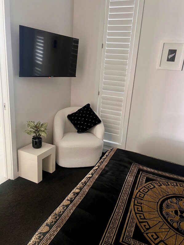 A modern bedroom with a flat-screen TV mounted on the wall, a small white side table with a potted plant, a white armchair with a black pillow, and a black and gold patterned bedspread. - The Whitianga Waterways Boathouse   (Whitianga)
