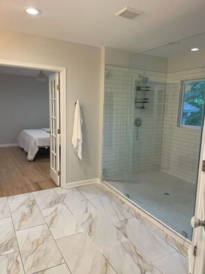 A modern bathroom with a glass-enclosed shower, marble flooring, and a visible bed in the adjacent room.