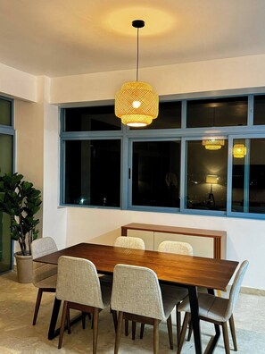 Un comedor con una mesa de madera y seis sillas, una lámpara colgante de fibras naturales y ventanas grandes con marcos azules.
