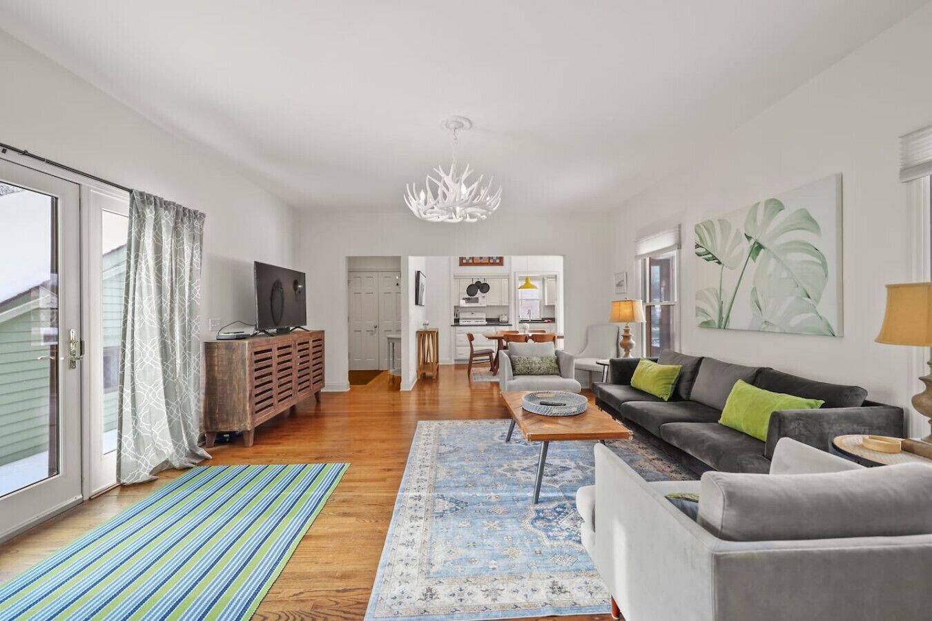 Una sala de estar moderna con un sofá, una mesa de centro y un televisor sobre un mueble de madera. Se observa una cocina al fondo, con armarios blancos y un comedor.