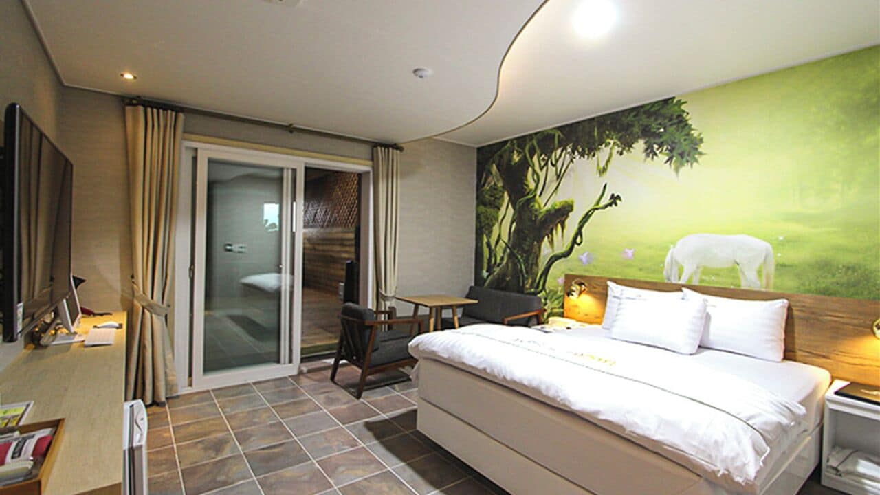 A hotel room with a large bed, a desk, a chair, a small table, a nightstand, and a sliding glass door leading to a balcony.