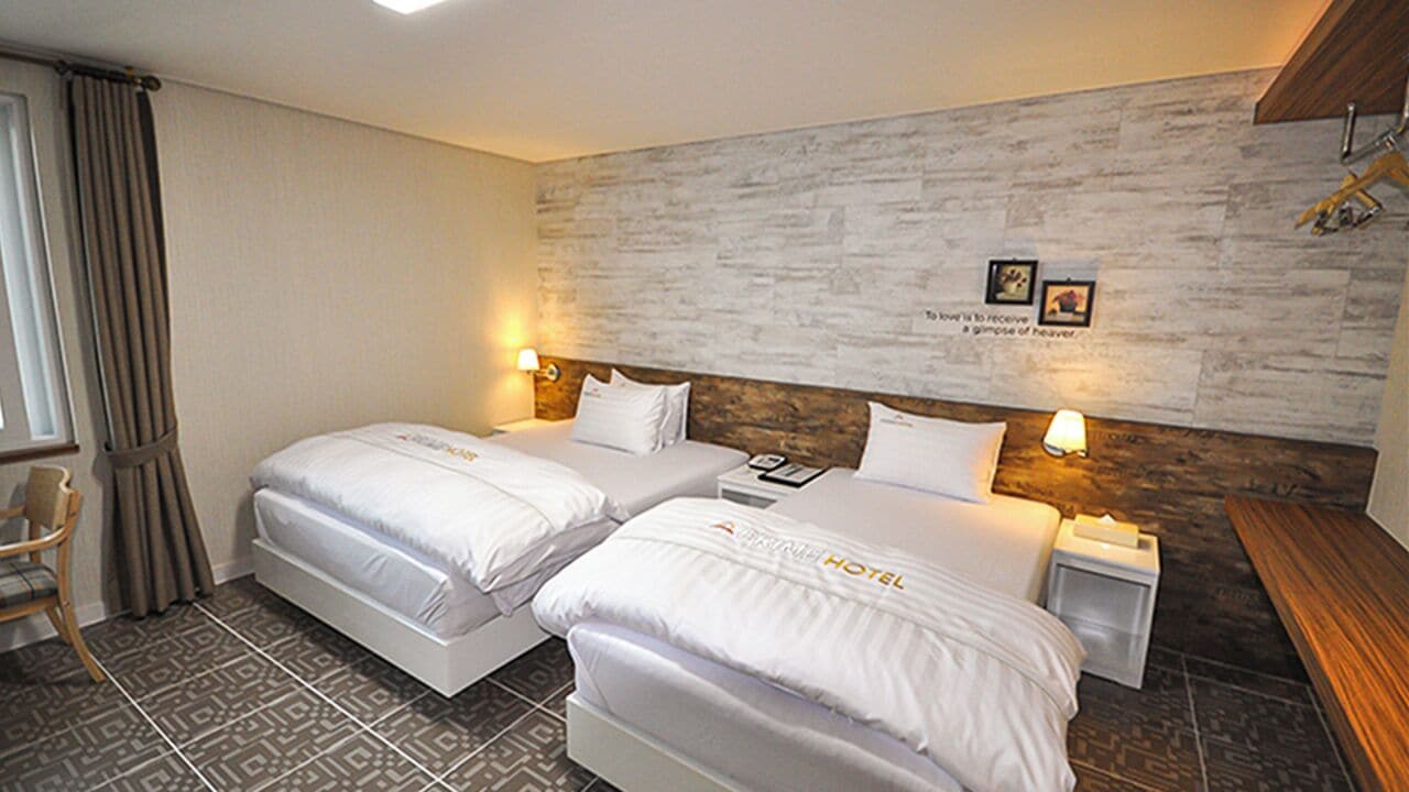 A hotel room with two beds, a wooden headboard, a small bedside table with a lamp, and a chair.