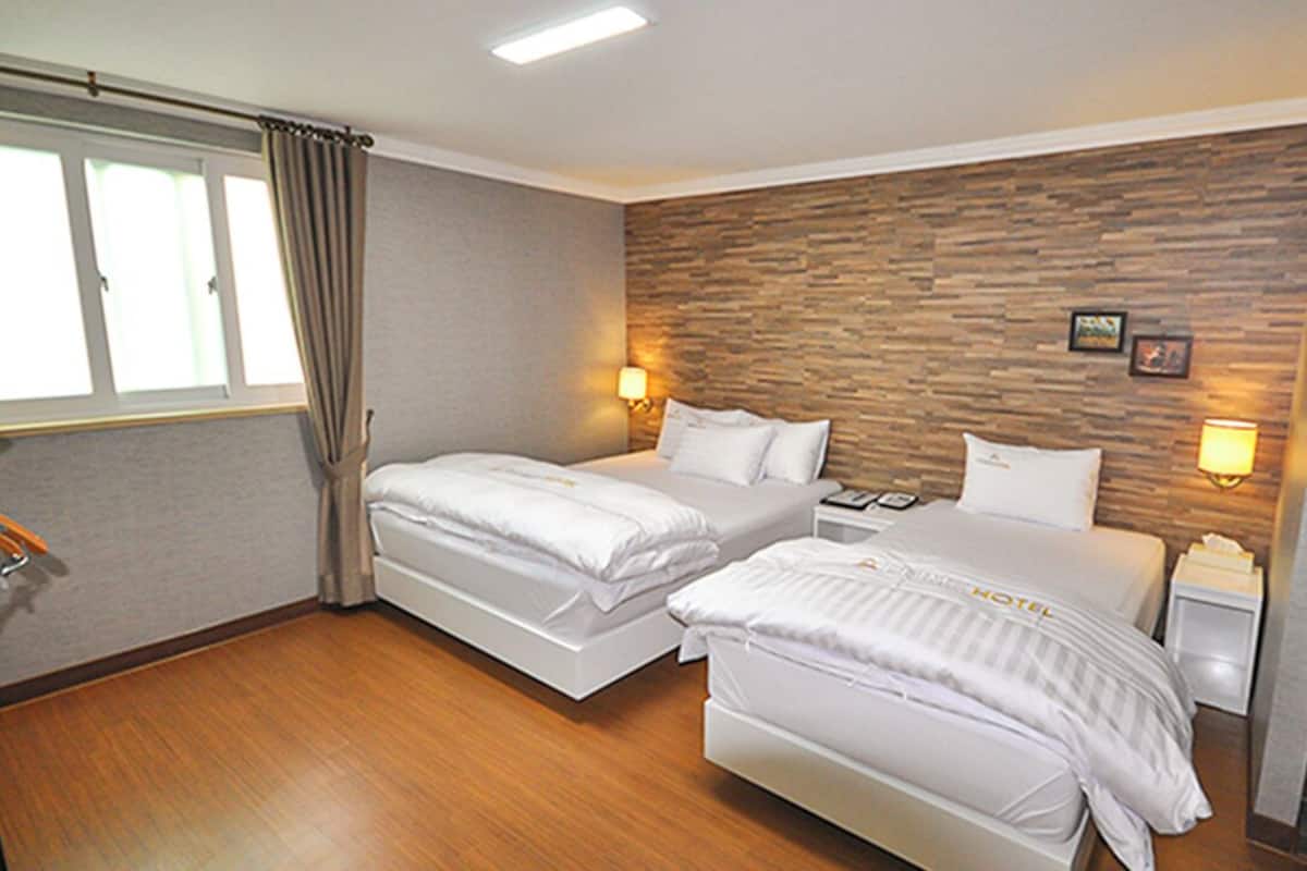 A hotel room with two beds, a wooden accent wall, bedside lamps, a window with curtains, and a small desk with a chair.