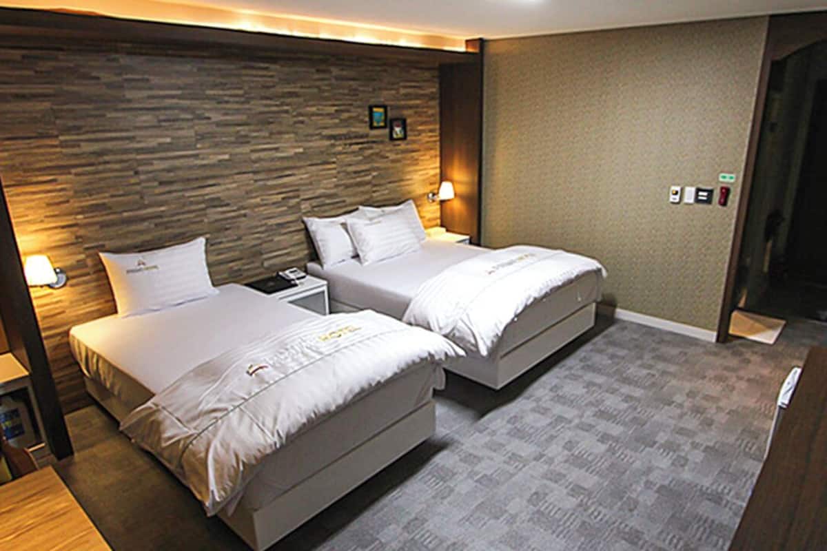 A hotel room with two beds, a wooden headboard, a carpeted floor, a desk with a chair, and a door.
