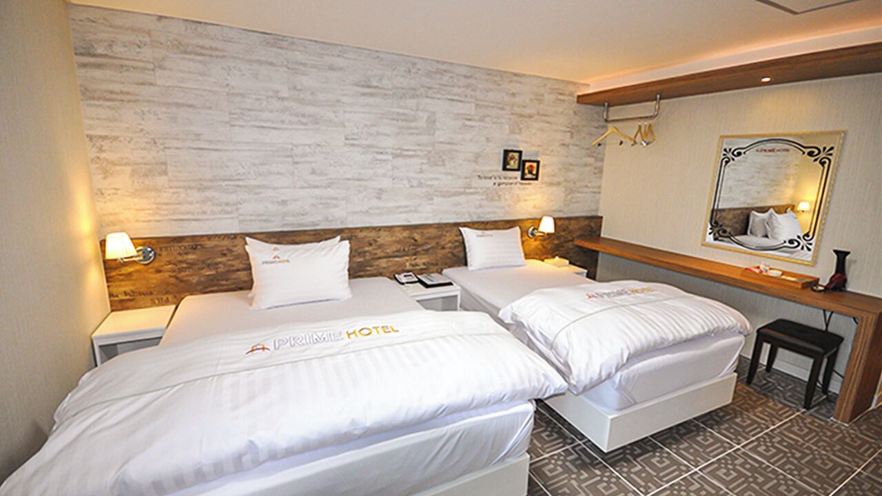 Una habitació d'hotel amb dos llits, un capçal de llit de fusta, una tauleta de nit amb una làmpada, un mirall i una taula petita.