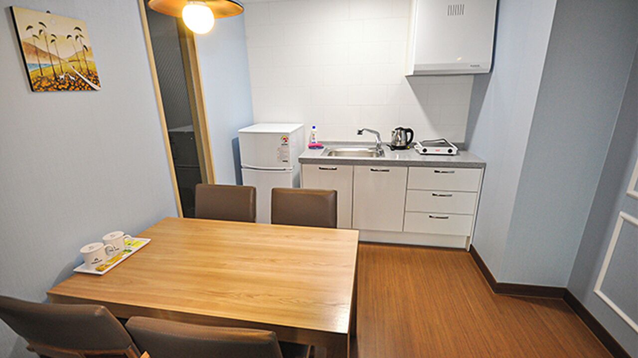 A compact kitchen with a dining area, including a wooden table and chairs, a refrigerator, a sink, and a kettle.