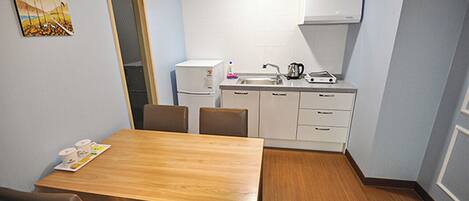 A compact kitchen with a dining area, including a wooden table and chairs, a refrigerator, a sink, and a kettle.